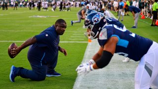 Titans defensive line coach Terrell Williams mimics a snap with a football while kneeling on the field prior to a game. Two Titans players in blue helmets and jerseys are standing up from their crouched stances to mimic their actions at the snap.